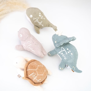 Könnte beinhalten: Vier Plüschtiere: ein grüner Wal mit gesticktem "Andre", ein rosa Seehund mit "Elena", ein blauer Dinosaurier mit "Jove" und eine beige Schildkröte mit "Patrik". Jedes Spielzeug hat ein einzigartiges Design und Farbschema.