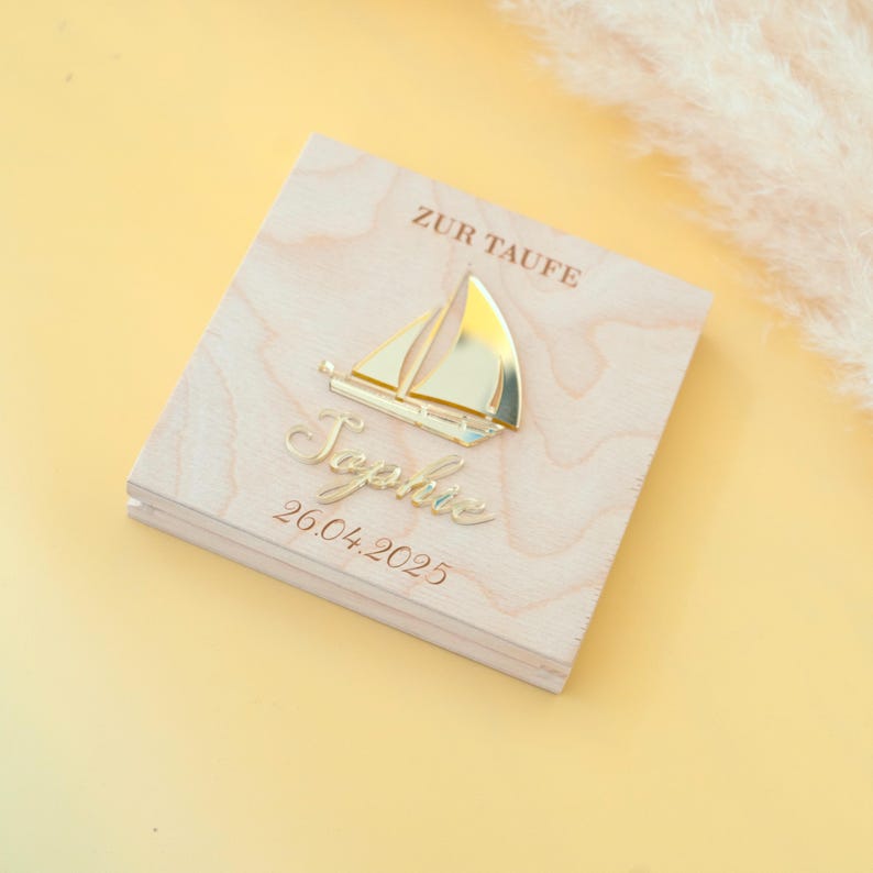 May include: A wooden box with a gold sailboat and the name "Sophie" engraved on it. The box also has the text "Zur Taufe" and the date "26.04.2025" engraved on it.