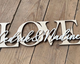 Könnte beinhalten: Ein Holzschild mit dem ausgeschnittenen Wort "LOVE" in der Mitte. Die Namen "Cedrik & Nadine" sind in weißer Kursivschrift innerhalb der Buchstaben geschrieben.