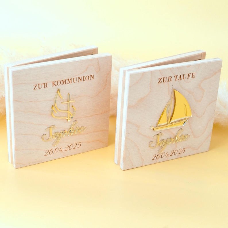 May include: Two wooden boxes with gold accents. The box on the left has a cross and flame design and the text "Zur Kommunion" and "26.04.2025". The box on the right has a sailboat design and the text "Zur Taufe" and "26.04.2025". Both boxes have the name "Sophie" written in cursive.