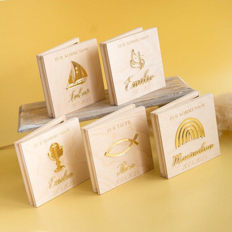 May include: Five wooden boxes with gold accents and engraved text. Each box features a different religious symbol, including a sailboat, a cross, a chalice, a fish, and a rainbow. The boxes are labeled with the names "Lukas", "Emilia", "Emilia", "Theo", and "Maximilian" and the dates of their respective events.