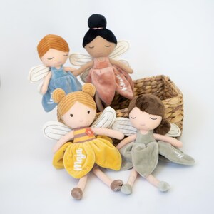 以下が含まれることがあります： 刺繍された顔立ちと布製の羽を持つ4体のぬいぐるみ妖精人形。各人形は青、ダスティピンク、マスタードイエロー、セージグリーンのベルベットのドレスを着ています。ドレスの花びらには白い刺繍で名前が入れられています。