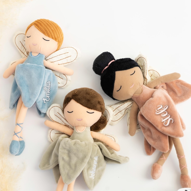 以下が含まれることがあります： 刺繍入りの名前が付いた3体のぬいぐるみ妖精人形。1体は赤毛でエミリアと書かれた青いドレス、1体は茶色の髪でヨハンナと書かれたセージグリーンのドレス、もう1体は黒髪をお団子にまとめステフィと書かれたダスティローズのドレスを着ています。