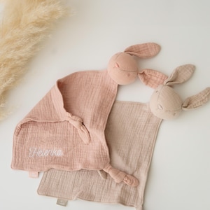 May include: Two baby comforters with bunny heads. One is pale pink with the name "Helenka" embroidered in white. The other is a light beige colour. Both have knotted corners and are made of soft, textured fabric.
