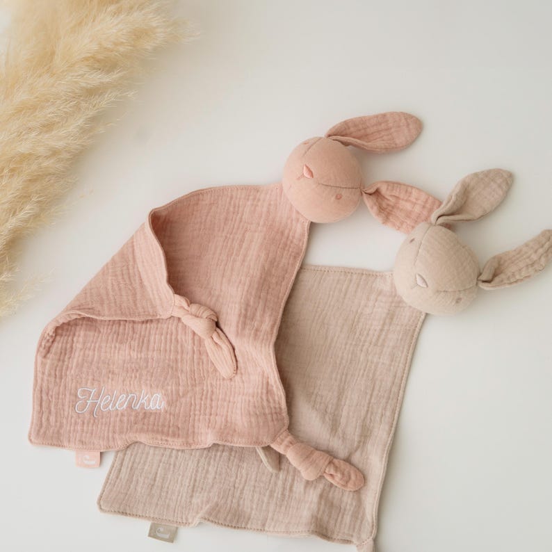May include: Two baby comforters with bunny heads. One is pale pink with the name "Helenka" embroidered in white. The other is a light beige colour. Both have knotted corners and are made of soft, textured fabric.