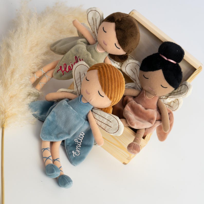 以下が含まれることがあります： 刺繍入りの羽と閉じた目を持つ3体のぬいぐるみ妖精人形。1体は茶色の髪でHeidiという名前入りの緑色のドレス、1体は赤毛でEmiliaという名前入りの青いドレス、もう1体は黒髪でピンクのドレスを着ています。