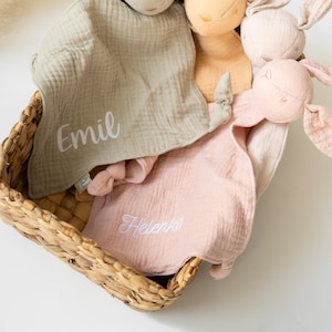 May include: A wicker basket contains several soft bunny toys and muslin security blankets. The blankets are in green and pink, with embroidered names. The bunnies are in beige, pink, and cream colours.