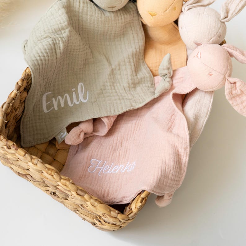 May include: A wicker basket contains several soft bunny toys and muslin security blankets. The blankets are in green and pink, with embroidered names. The bunnies are in beige, pink, and cream colours.