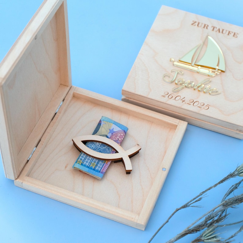 May include: A wooden box with a hinged lid, engraved with the words "Zur Taufe" and a sailboat. The box is open, revealing a wooden fish-shaped ornament and a folded bill inside.