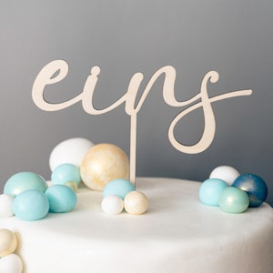 Op de afbeelding: Een witte taart versierd met blauwe en gouden ronde snoepjes en een houten taarttopper met de tekst "eins".