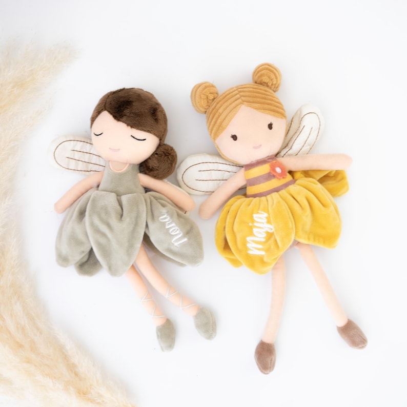 以下が含まれることがあります： 刺繍された顔立ちと翼を持つ2体の妖精のぬいぐるみ。左のぬいぐるみは茶色の髪で、Noraと刺繍されたセージグリーンのドレスを着ています。右のぬいぐるみはブロンドの髪で、ストライプのボディスとMajaと刺繍された黄色いドレスを着ています。