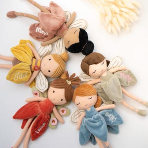 Könnte beinhalten: Fünf Plüsch-Feenpuppen mit gestickten Namen auf ihren Samtröcken. Jede Puppe hat aufgestickte Gesichtszüge, Stoffflügel und lange Gliedmaßen. Die Puppen sind in verschiedenen Farben gekleidet, darunter Altrosa, Gelb, Braun, Salbeigrün und Blau.