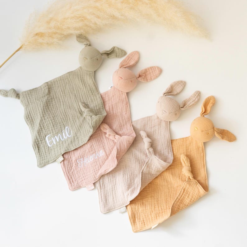 May include: Four pastel-coloured baby security blankets with bunny heads. Each blanket is a different colour: sage green, light pink, beige, and peach. The sage green and light pink blankets have embroidered names. The blankets are made of a soft, textured fabric.