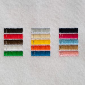 以下が含まれることがあります： 3つの垂直な列に配置された15個の長方形の刺繍糸のサンプルを含むカラーチャート。各サンプルには1から15までの白い番号が付けられており、黒、赤、茶色、白、緑、灰色、ベージュ、黄色、オレンジ、青、ピンク、ティールなど、さまざまな色が表示されています。