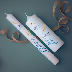 Könnte beinhalten: Zwei weiße Kerzen mit blauen und goldenen Designs. Die Kerzen sind mit Weizenähren, Fischen und dem Text "Nathalie 31.01.2023" und "Kommt, ißt und Esst!" verziert.