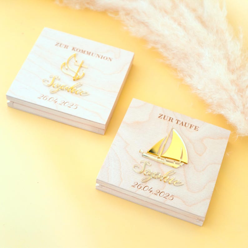 May include: Two wooden boxes with gold accents. The top box has an anchor and the text "Zur Kommunion" and "26.04.2025". The bottom box has a sailboat and the text "Zur Taufe" and "26.04.2025".