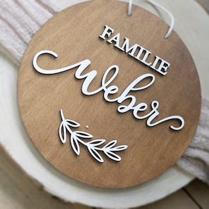 Könnte beinhalten: Ein rundes Holzschild mit dem Text "Familie Weber" in weißen Buchstaben. Das Schild hat unten ein weißes Zweigdesign.