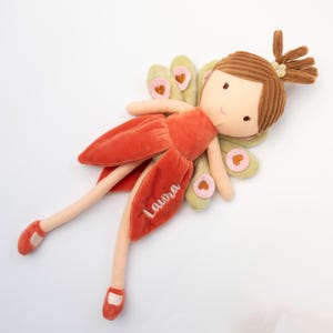 以下が含まれることがあります： 茶色のコーデュロイの髪をトップノットに結び、Lauraという名前が刺繍された錆びたオレンジ色のベルベットのドレスを着た、ぬいぐるみのような妖精の人形。ピンクのハートのアクセントが付いた淡い緑色の羽と、長く細い手足、お揃いの靴が特徴です。