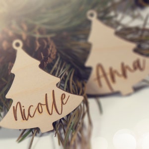 May include: Two wooden Christmas tree ornaments with the names "Nicole" and "Anna" engraved on them. The ornaments are lying on a bed of pine needles.