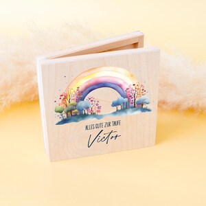 Peut inclure: Un coffret en bois avec un motif arc-en-ciel &agrave; l'aquarelle. L'arc-en-ciel surplombe un paysage d'arbres et de nuages. Le texte "ALLES GUTE ZUR TAUFE" et le nom "Victor" sont &eacute;crits en dessous.