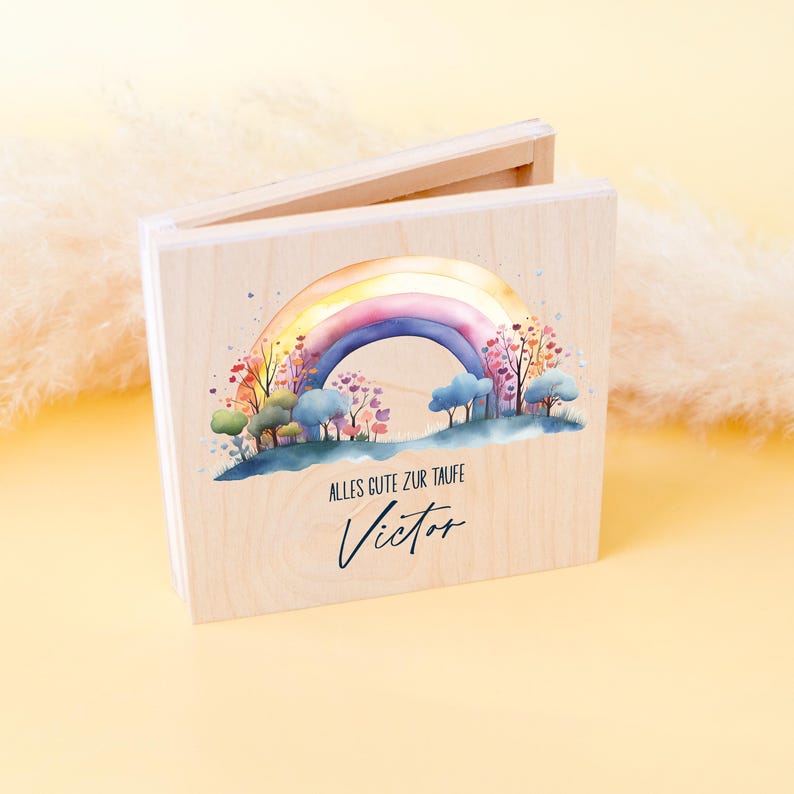 Peut inclure: Un coffret en bois avec un motif arc-en-ciel &agrave; l'aquarelle. L'arc-en-ciel surplombe un paysage d'arbres et de nuages. Le texte "ALLES GUTE ZUR TAUFE" et le nom "Victor" sont &eacute;crits en dessous.