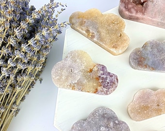 May include: A set of seven cloud-shaped amethyst crystal carvings in various shades of purple, pink, and yellow. The crystals are arranged on a white surface.