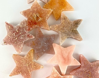 May include: A collection of nine star-shaped soap bars in various shades of pink, orange, and brown. The soap bars have a speckled appearance and are arranged in a cluster on a white surface.