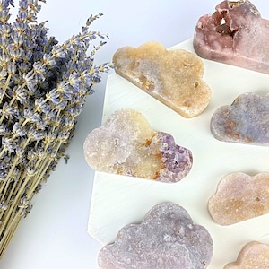 May include: A set of seven cloud-shaped amethyst crystal carvings in various shades of purple, pink, and yellow. The crystals are arranged on a white surface.