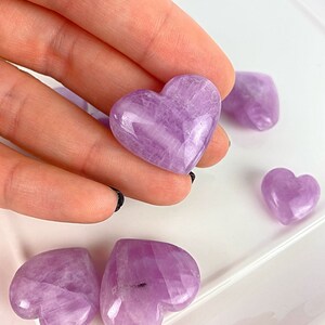 May include: A collection of polished amethyst heart-shaped stones. The stones are a light purple colour and have a smooth, polished finish.