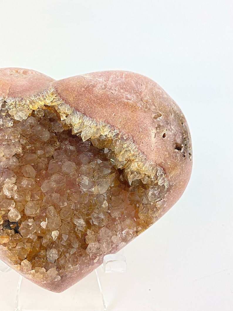 May include: A heart-shaped geode with a pink exterior and a cavity filled with clear quartz crystals. The crystals are clustered together and have a light brown color.