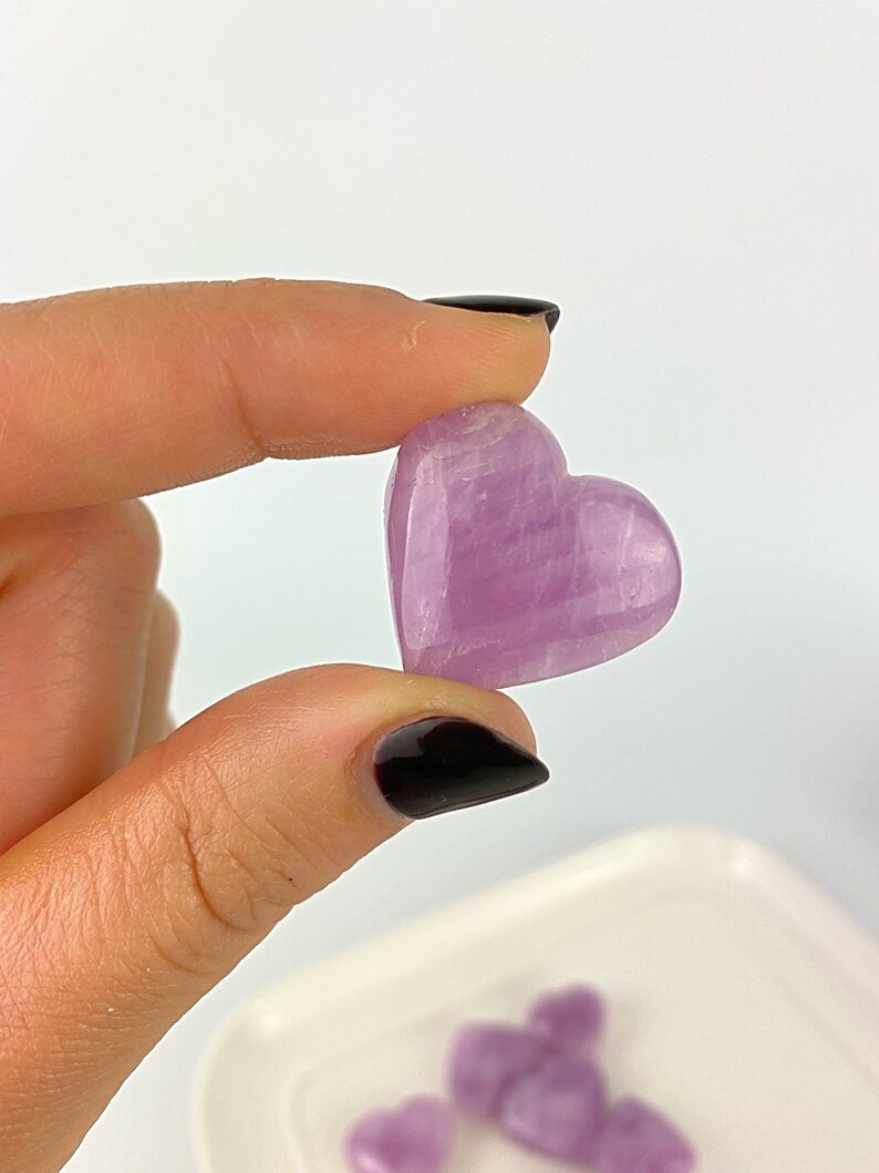 May include: A small, heart-shaped, polished amethyst crystal with a light purple hue. The crystal has a smooth, polished surface and is a light purple colour.
