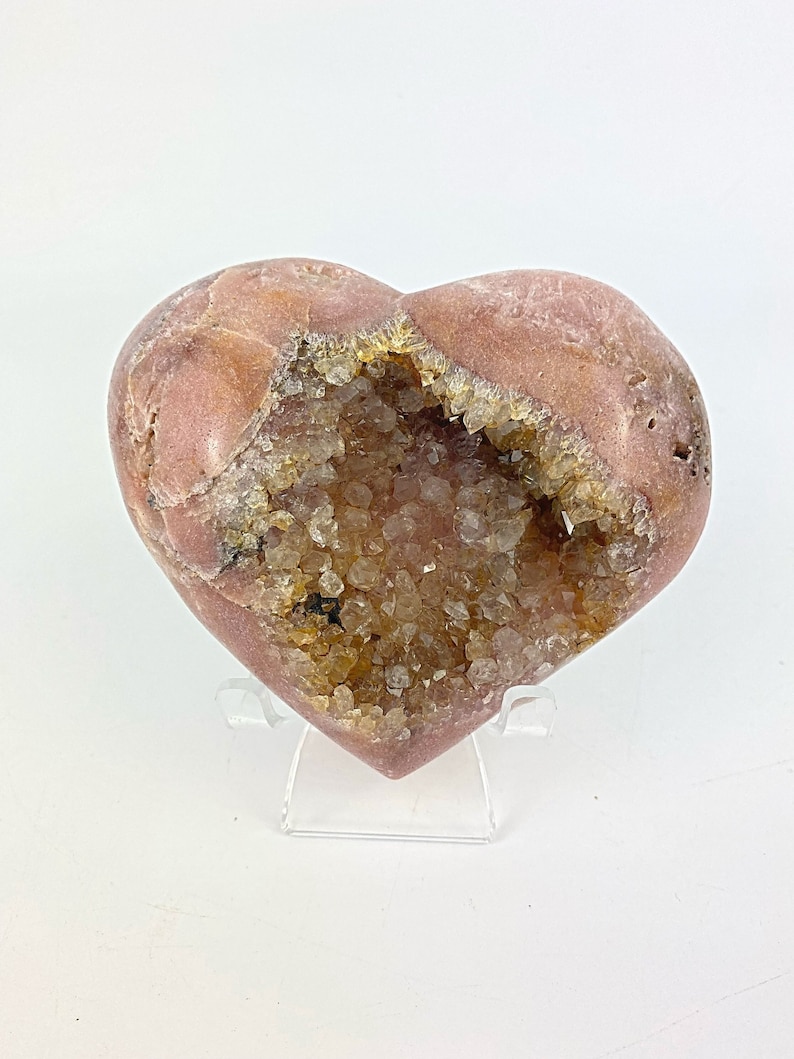 May include: A heart-shaped geode with a pink exterior and a clear quartz crystal interior. The geode is on a clear acrylic stand.