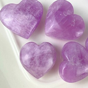 May include: Five purple heart-shaped crystals on a white surface. The crystals are smooth and polished.