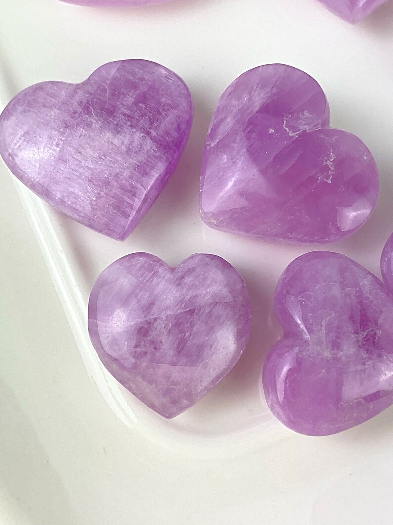 May include: Five purple heart-shaped crystals on a white surface. The crystals are smooth and polished.