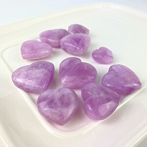 May include: A collection of eight purple heart-shaped stones on a white plate. The stones are smooth and polished, with a light purple hue.