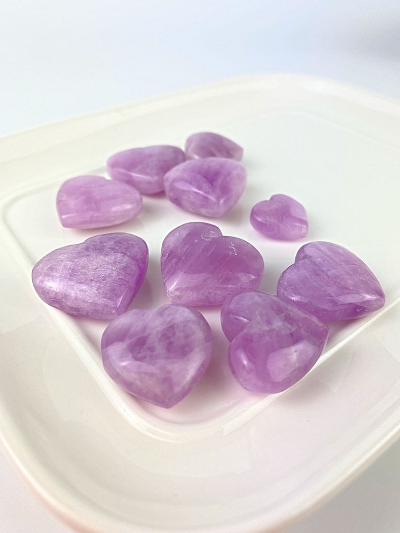 May include: A collection of eight purple heart-shaped stones on a white plate. The stones are smooth and polished, with a light purple hue.