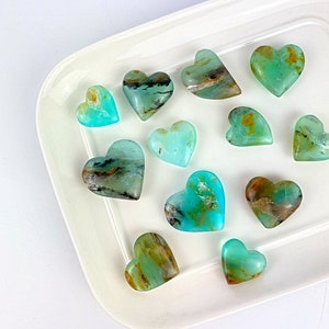 Puede incluir: Una colección de piedras preciosas en forma de corazón en tonos turquesa, verde azulado y marrón, dispuestas sobre una fuente rectangular blanca. Las piedras pulidas varían en tamaño y muestran patrones únicos, creando una estética natural.