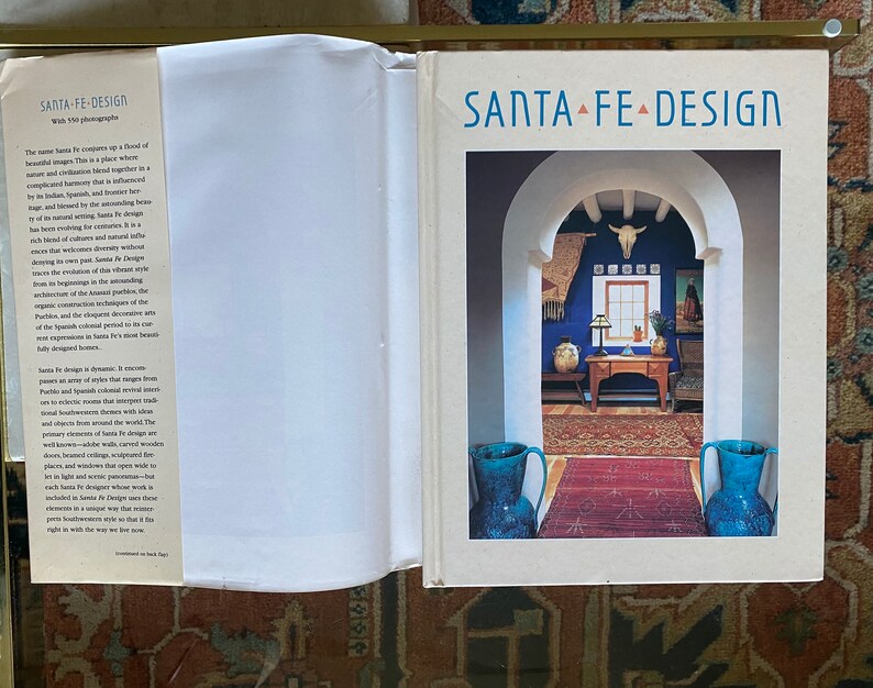 May include: A book cover with the title "Santa Fe Design" in a white font on a white background. The cover features a photograph of a doorway with a blue wall and a rug on the floor. There are two blue vases on either side of the doorway.