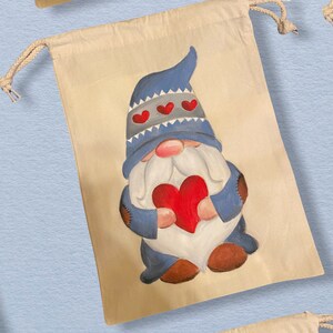 Può includere: Una borsa beige con coulisse con un gnomo dipinto che tiene un cuore rosso. Lo gnomo indossa un cappello blu con un bordo bianco e cuori rossi.