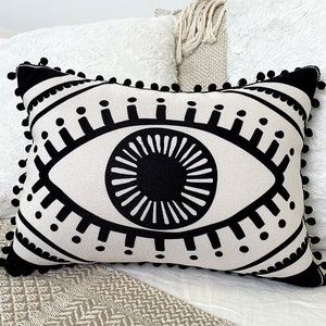 May include: A white throw pillow with a black eye design. The eye is surrounded by a black circle with a white center. The pillow has a black pom-pom trim.