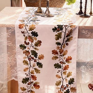 May include: A white table runner featuring two vertical, symmetrical botanical patterns of brown, green, and orange oak leaves and acorns. The runner is placed on a table covered with a white lace tablecloth, with metallic candle holders visible.