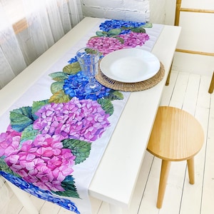 Könnte beinhalten: Ein weißer Tischläufer mit einem Blumendesign aus rosa, blauen und grünen Hortensien. Der Läufer liegt auf einem weißen Tisch mit einem weißen Teller und einem Glas auf einem geflochtenen Untersetzer.