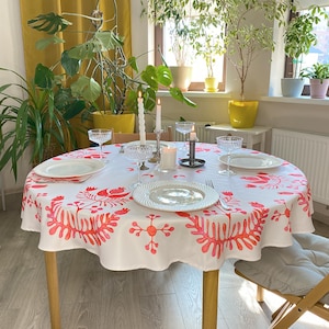 Pode incluir: Uma toalha de mesa redonda branca com um padrão floral vermelho cobre uma mesa de madeira montada para uma refeição. A mesa é rodeada por uma cadeira de madeira e decorada com pratos, talheres e taças de cristal. Velas acesas contribuem para o ambiente.