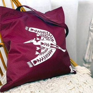 May include: A maroon tote bag with a white design of a stylized bird with floral accents.