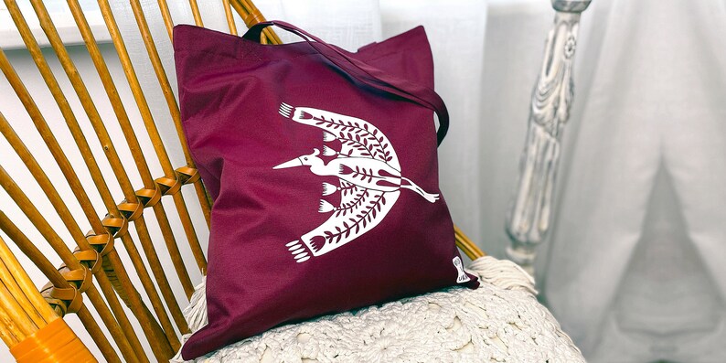 May include: A maroon tote bag with a white design of a stylized bird with floral accents.