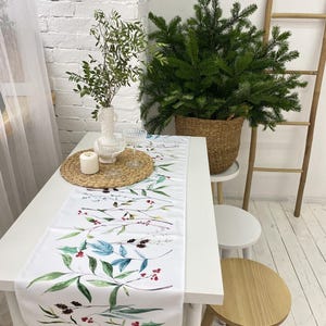 Puede incluir: Camino de mesa blanco con un diseño botánico en acuarela con hojas verdes, detalles azules y bayas rojas. El camino de mesa está colocado sobre una mesa blanca, con un pequeño árbol de Navidad al fondo. También hay una vela y cristalería sobre la mesa.