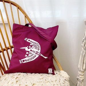 May include: A burgundy tote bag with a white design of a stylized bird with floral accents. The bag is sitting on a wicker chair.