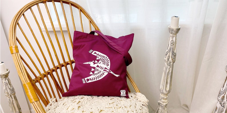 May include: A burgundy tote bag with a white design of a stylized bird with floral accents. The bag is sitting on a wicker chair.
