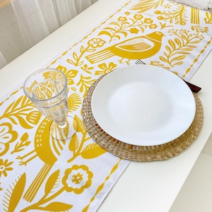 May include: A white table runner with a yellow floral pattern featuring a bird design. The runner is set with a white plate and a glass.