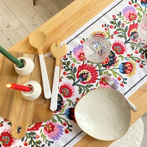 Könnte beinhalten: Eine Tischdekoration mit einem weißen, floralen Tischläufer mit roten, rosa und blauen Blumen. Enthält hölzerne Salatbestecke, einen weißen Teller, klares Glasgeschirr und Kerzen in weißen Haltern.